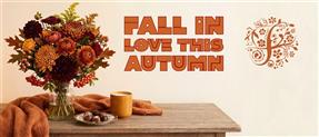 Warm autumn floral arrangement in a glass vase alongside a candle and chocolates on a rustic wooden table, set against a cream background